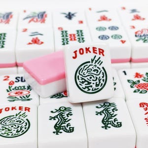 Pu&ograve; includere: Una collezione di tessere del Mahjong bianche con retro rosa, caratterizzate da vari disegni incisi in verde e rosso, tra cui draghi, fiori e la parola JOKER stampata in rosso sopra un grafico circolare sulla tessera in primo piano.