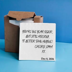 May include: A white square paper napkin featuring black printed text that reads: You're one year older, but still rocking, it better than anyone! Cheers, Emma XX, and the date Dec 8, 2026. The napkin is positioned in front of an open brown cardboard box.