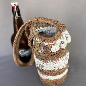 May include: A woven bottle carrier in shades of brown, white, and red, with a braided handle. The carrier is designed to hold a bottle, with a brown glass bottle visible in the background.