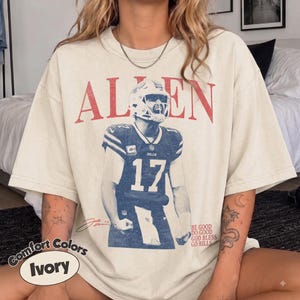 May include: An ivory t-shirt featuring a large red graphic of the name ALLEN above a blue-toned image of a football player wearing a jersey with the number 17. The text BE GOOD DO GOOD GOD BLESS GO BILLS is printed in small red letters on the lower right.