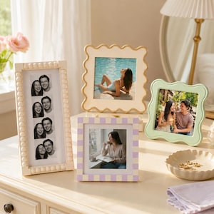 May include: A collection of four picture frames: a tall rectangular cream frame with beaded edges, a square cream frame with wavy edges, a square light green frame with wavy edges, and a square frame with a light purple and cream checkerboard pattern.