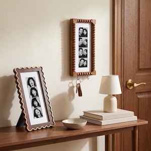 Puede incluir: Un marco de fotos rectangular de madera con un borde dorado ondulado que contiene una tira vertical de cuatro fotos en blanco y negro, y un marco de pared de madera con cuentas y dos ganchos metálicos que sostienen un juego de llaves plateadas.