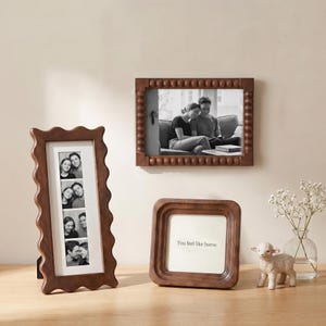 May include: Three brown picture frames of varying shapes and sizes are displayed on a light-colored surface. One frame has a wavy edge and holds four black and white photos. Another frame, square with rounded corners, has the text "You feel like home." The third frame is rectangular with a beaded border.