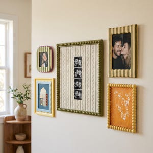 May include: A collection of five wall-mounted picture frames featuring various designs, including olive green beaded frames, a yellow scalloped frame, and a striped frame, displaying black and white photographs and floral artwork on a light wall.
