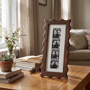 Peut inclure: Un cadre photo en bois marron avec un bord ondulé, contenant une série de photos de style photomaton en noir et blanc. Le cadre est posé sur une table en bois. Une petite plante en pot et un vase de fleurs sont également sur la table.