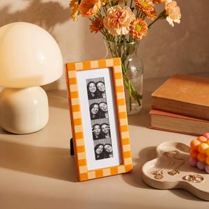 May include: A rectangular picture frame with an orange and cream checkered pattern containing a vertical black and white photo strip of two people. The frame stands upright on a surface next to a lamp, a vase of flowers, books, and a trinket tray.