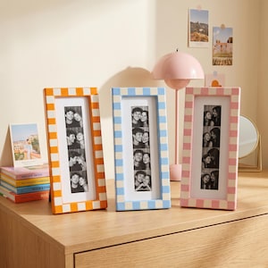 May include: Three rectangular picture frames with a chequered pattern in orange, light blue, and pink. Each frame holds a vertical black and white photo strip featuring four stacked portraits of two people. The frames are standing upright.