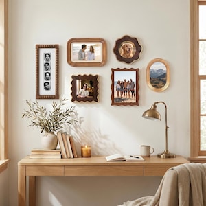 Può includere: Una collezione di foto incorniciate in varie forme e dimensioni, montate su una parete bianca. Le cornici sono principalmente marroni con diversi bordi decorativi. Una scrivania in legno con libri, una candela e una lampada è in primo piano.