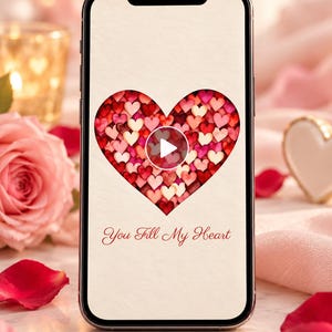 May include: A smartphone screen displays a large heart shape filled with numerous small red, pink, and white hearts. A white play button icon is centered within the heart. Below the heart, the text reads You Fill My Heart in red script.