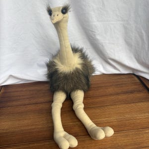 May include: A plush ostrich toy featuring a long, cream-colored neck and head with dark, expressive eyes and prominent eyelashes. The body is covered in dark brown, shaggy faux fur, and the long, slender legs are cream-colored with two-toed feet.
