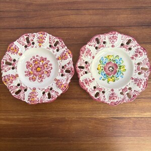 May include: Two round ceramic plates with scalloped, pierced edges. The left plate features a central pink floral motif with yellow accents. The right plate displays a central flower with pink, blue, and yellow petals. Both have pink floral borders.