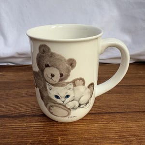 May include: A cream-colored ceramic mug featuring a detailed illustration of a brown teddy bear hugging a small white kitten with bright blue eyes. The artist signature V. Miller is printed in small black text near the base of the illustration.