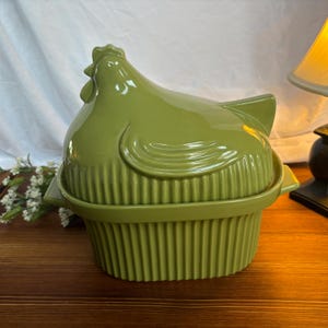 May include: A green ceramic chicken-shaped casserole dish. The lid is shaped like a chicken, and the base has vertical ridges. The dish is designed for cooking and serving food. The color is a muted, olive green.