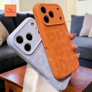May include: Two phone cases, one orange with a leopard print design and the other light purple. The orange case is held up, revealing the camera lenses. The cases are on a wooden surface.