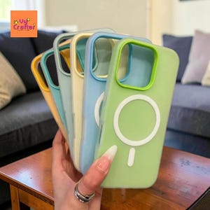 May include: A hand holds a stack of colorful phone cases. The cases are in shades of yellow, teal, light blue, and green. Each case has a white circle and a small line on the back. The cases are made of a soft, flexible material.