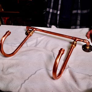 May include: A wall-mounted rack constructed from polished copper pipes and brass fittings. The structure features two large, curved hooks extending outward from a horizontal bar, which is secured to the wall by two circular black metal flanges.