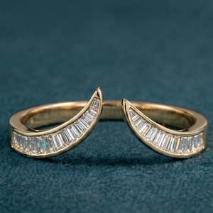 Könnte beinhalten: Ein goldfarbener Ring mit geteiltem Schaftdesign. Jede Seite des Rings hat eine gebogene Reihe rechteckig geschliffener, klarer Edelsteine. Der Ring ist vor einem dunkelblauen Hintergrund platziert, der das Funkeln der Steine hervorhebt.