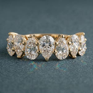 Op de afbeelding: Een gouden ring met zeven peervormige diamanten. De diamanten zijn dicht op elkaar geplaatst, waardoor een continu sprankelend effect ontstaat. De ring is geplaatst op een teal achtergrond, die de schittering van de edelstenen benadrukt.