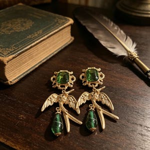 May include: A pair of gold-toned earrings featuring a rectangular emerald green faceted stone at the top, connected to a detailed gold bird motif with outstretched wings, and finished with a dangling teardrop-shaped green marbled bead.
