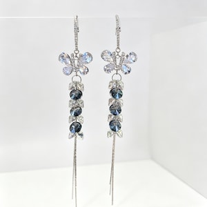 May include: A pair of silver-toned drop earrings featuring a butterfly-shaped charm encrusted with clear crystals. Below the butterfly, a vertical strand of alternating clear and dark grey faceted beads hangs, ending in two thin silver-toned chains.