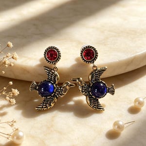 May include: A pair of drop earrings featuring a gold-toned metal bird design with textured wings and a round, deep blue cabochon stone at the center. Each earring is topped with a circular, faceted red crystal stud set in a twisted metal frame.