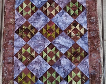 May include: A quilt with a brown and purple diamond pattern. The quilt is made up of smaller squares with a variety of colors, including green, brown, and white. The quilt is edged with a brown border.