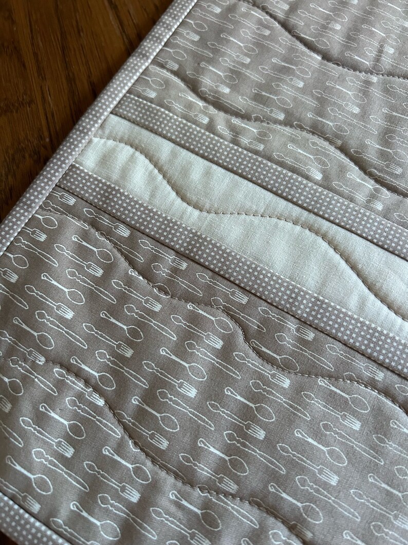 K&ouml;nnte beinhalten: Nahaufnahme einer braunen, gesteppten K&uuml;chenmatte mit einem wei&szlig;en Muster aus L&ouml;ffeln und Gabeln. Die Matte hat einen wei&szlig;en Rand und ein gestepptes Design.