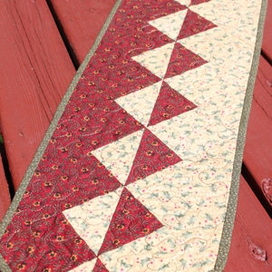 May include: A red and cream quilted table runner with a geometric pattern of triangles. The runner has a green border and is made of fabric with a floral print.