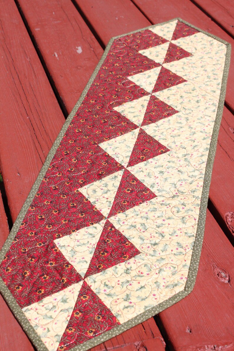 May include: A red and cream quilted table runner with a geometric pattern of triangles. The runner has a green border and is made of fabric with a floral print.