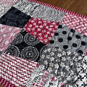 K&ouml;nnte beinhalten: Ein Patchwork-Deckenst&uuml;ck mit verschiedenen Mustern in Schwarz, Wei&szlig;, Grau und Rosa. Die Decke besteht aus Quadraten mit unterschiedlichen Designs, darunter Punkte, Blumen und geometrische Formen. Die Decke ist mit einem roten und wei&szlig;en Rand versehen.