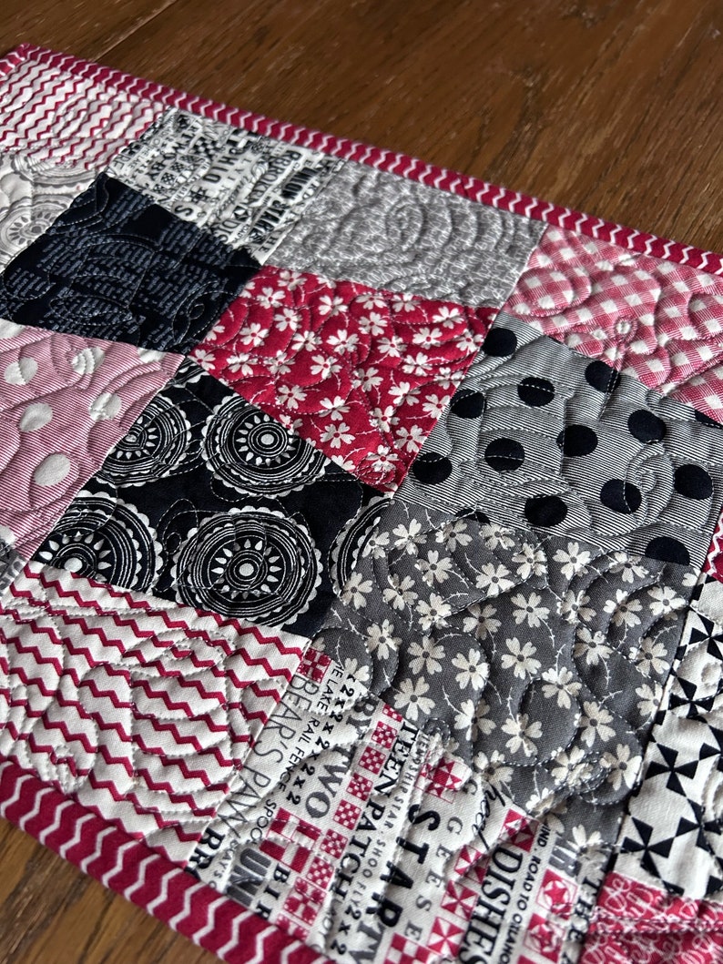 K&ouml;nnte beinhalten: Ein Patchwork-Deckenst&uuml;ck mit verschiedenen Mustern in Schwarz, Wei&szlig;, Grau und Rosa. Die Decke besteht aus Quadraten mit unterschiedlichen Designs, darunter Punkte, Blumen und geometrische Formen. Die Decke ist mit einem roten und wei&szlig;en Rand versehen.