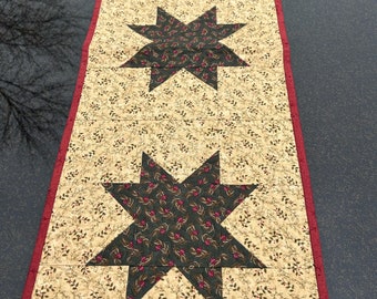 May include: A table runner with a red border and a beige background with a small floral pattern. Three large dark green stars with a smaller floral pattern are centered on the runner.