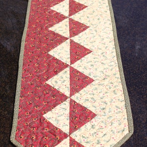 May include: A red and cream quilted table runner with a geometric pattern. The runner has a green border and is made of fabric with a floral print.