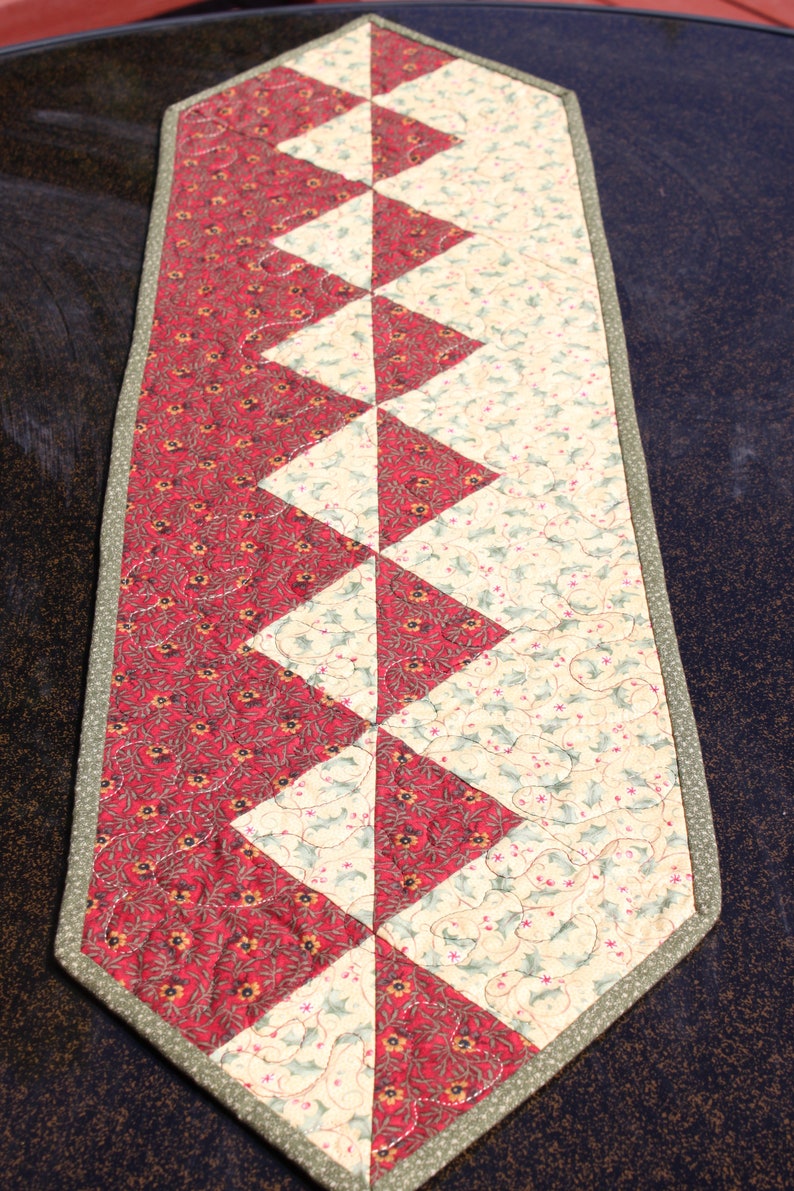 May include: A red and cream quilted table runner with a geometric pattern. The runner has a green border and is made of fabric with a floral print.