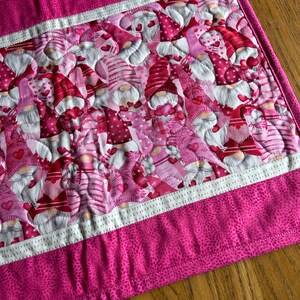 May include: Pink quilted table runner with a pattern of gnomes wearing pink and red hats and holding hearts. The runner has a white and gray trim.