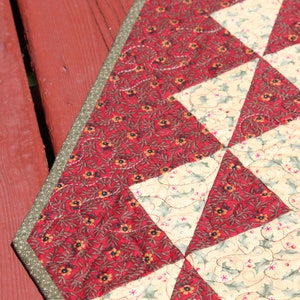 May include: A red and cream quilted table runner with a green border. The fabric features a floral pattern with small red flowers and green leaves.