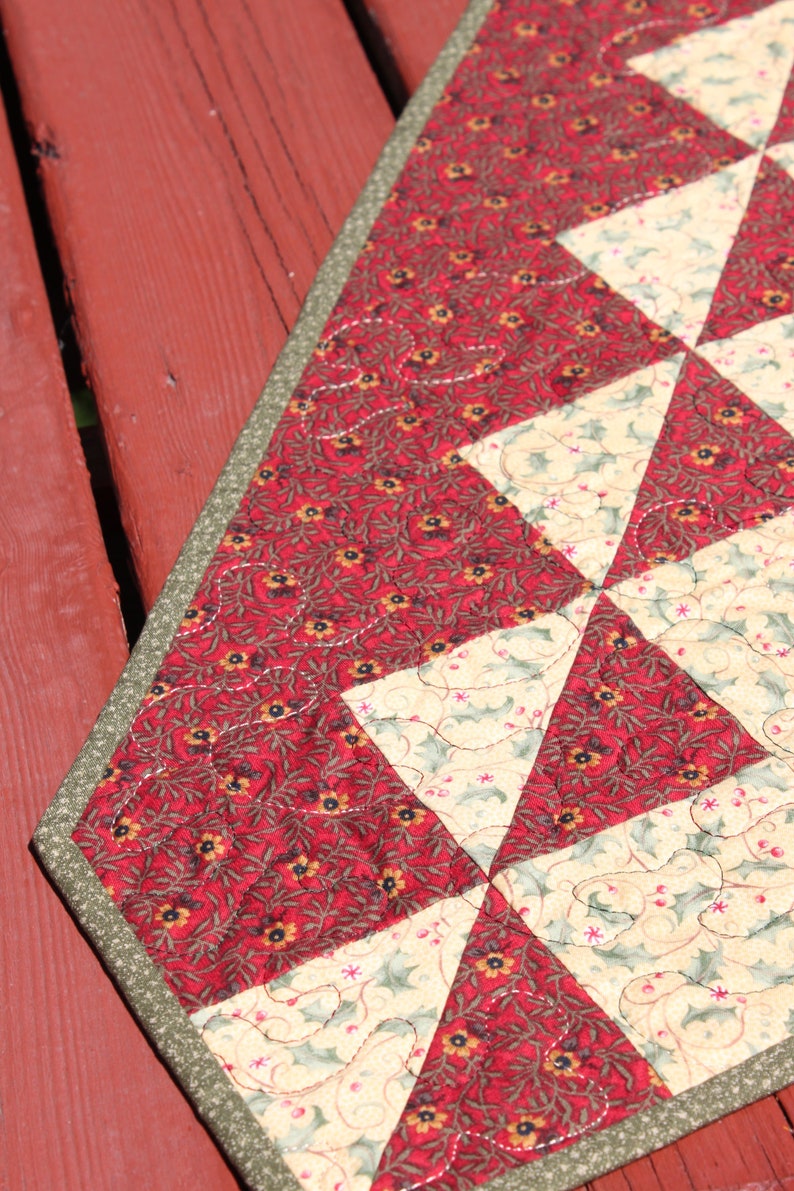May include: A red and cream quilted table runner with a green border. The fabric features a floral pattern with small red flowers and green leaves.