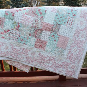 May include: A white quilt with a pink and blue patchwork design. The quilt is folded and hanging over a wooden railing.