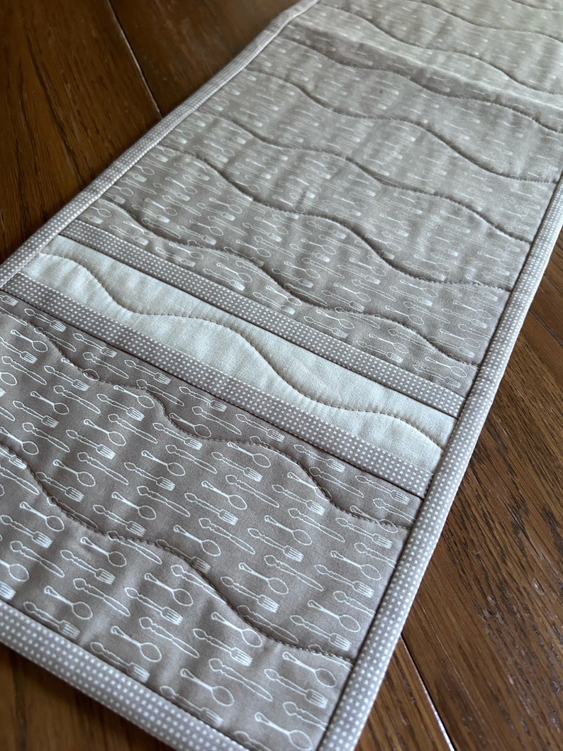 K&ouml;nnte beinhalten: Ein beiger Tischl&auml;ufer mit einem wei&szlig;en und beigen Muster aus L&ouml;ffeln und Gabeln. Der L&auml;ufer hat einen wei&szlig;en Rand mit einem kleinen wei&szlig;en Punktmuster.