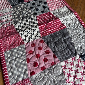 K&ouml;nnte beinhalten: Ein roter und grauer Patchwork-Tischl&auml;ufer mit gestepptem Design. Der L&auml;ufer zeigt verschiedene Muster, darunter Punkte, Gingham und florale Drucke.