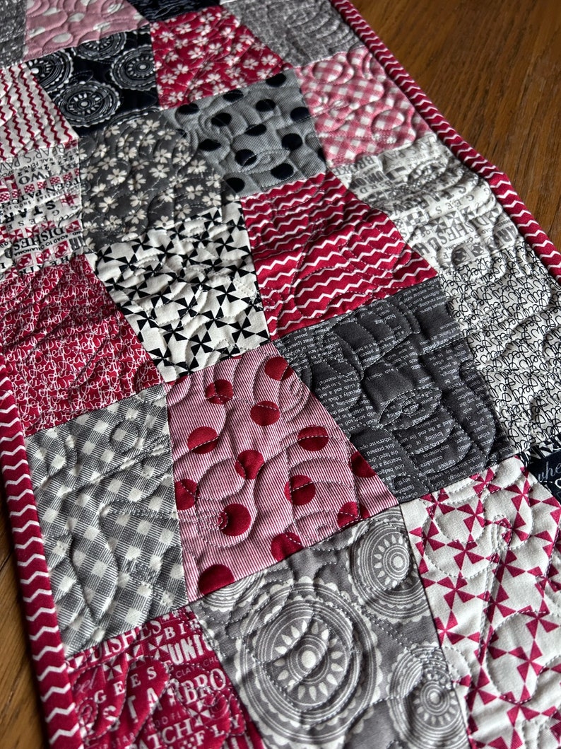 K&ouml;nnte beinhalten: Ein roter und grauer Patchwork-Tischl&auml;ufer mit gestepptem Design. Der L&auml;ufer zeigt verschiedene Muster, darunter Punkte, Gingham und florale Drucke.