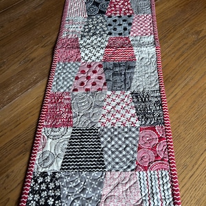 K&ouml;nnte beinhalten: Ein roter und grauer Patchwork-Tischl&auml;ufer mit einem wei&szlig;en Zickzack-Saum. Der L&auml;ufer besteht aus verschiedenen Stoffen mit unterschiedlichen Mustern, darunter Punkte, Streifen und Blumenmuster.