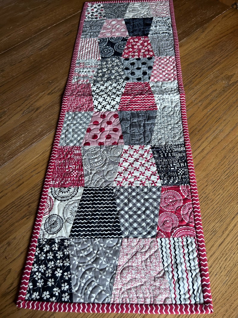 K&ouml;nnte beinhalten: Ein roter und grauer Patchwork-Tischl&auml;ufer mit einem wei&szlig;en Zickzack-Saum. Der L&auml;ufer besteht aus verschiedenen Stoffen mit unterschiedlichen Mustern, darunter Punkte, Streifen und Blumenmuster.