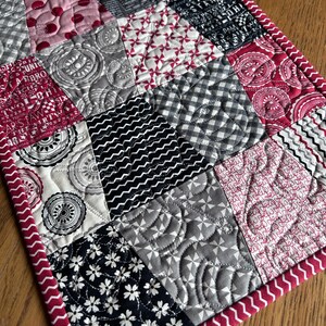 K&ouml;nnte beinhalten: Ein gesteppter Tischl&auml;ufer mit Patchwork-Design in verschiedenen schwarzen, wei&szlig;en und roten Mustern. Der L&auml;ufer hat einen roten Rand mit einem wei&szlig;en Zickzackmuster.