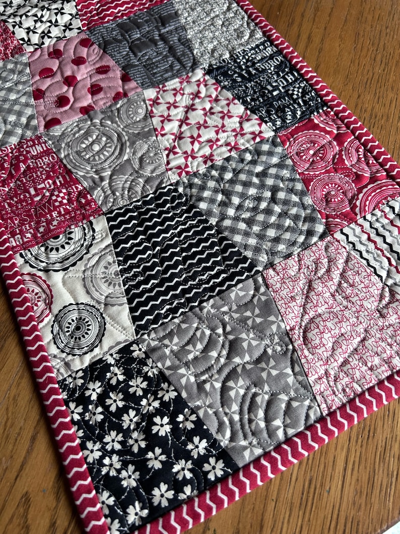 K&ouml;nnte beinhalten: Ein gesteppter Tischl&auml;ufer mit Patchwork-Design in verschiedenen schwarzen, wei&szlig;en und roten Mustern. Der L&auml;ufer hat einen roten Rand mit einem wei&szlig;en Zickzackmuster.