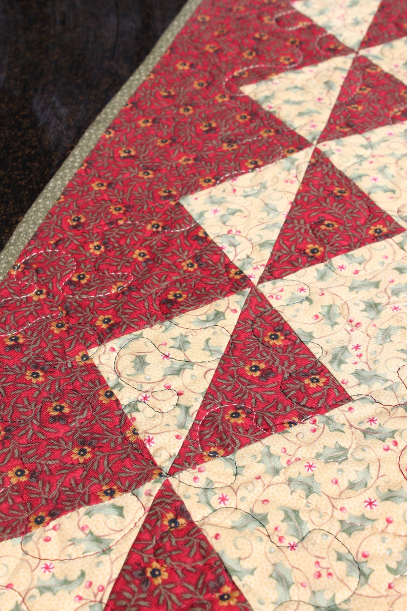 May include: A quilted table runner with a geometric pattern of red and cream triangles. The fabric features a floral print with small red flowers and green leaves.