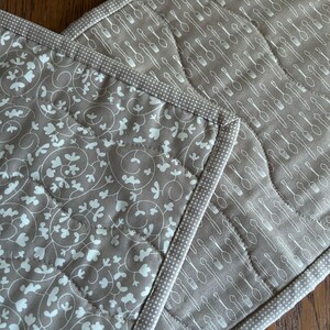 K&ouml;nnte beinhalten: Zwei braune, gesteppte Topflappen mit wei&szlig;en Designs. Ein Topflappen hat ein Muster aus Bl&auml;ttern und Ranken, der andere ein Muster aus L&ouml;ffeln und Gabeln.