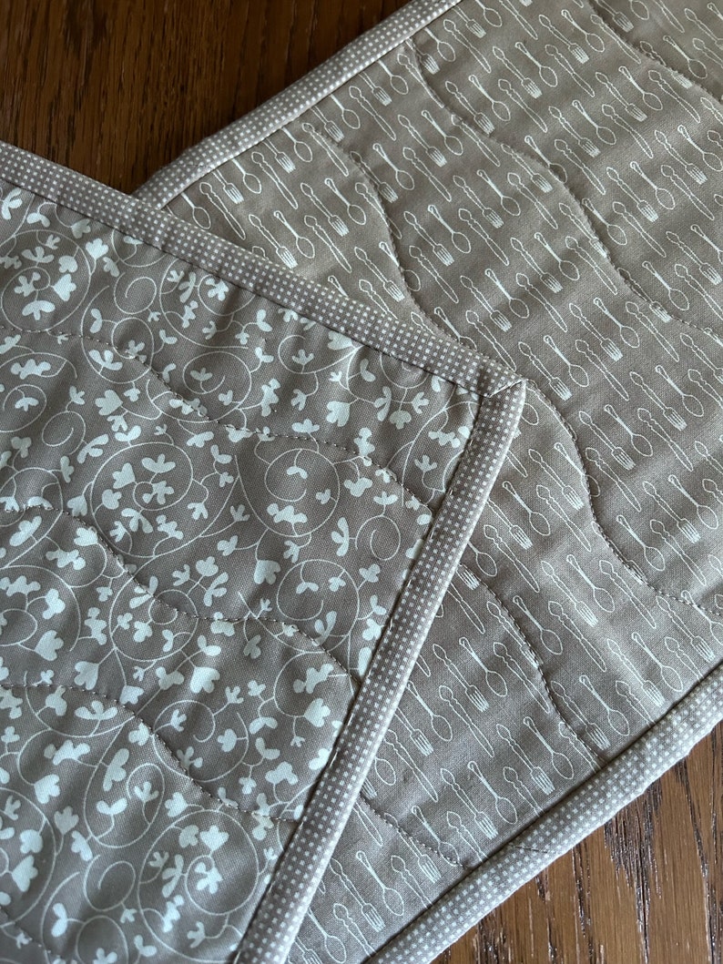K&ouml;nnte beinhalten: Zwei braune, gesteppte Topflappen mit wei&szlig;en Designs. Ein Topflappen hat ein Muster aus Bl&auml;ttern und Ranken, der andere ein Muster aus L&ouml;ffeln und Gabeln.