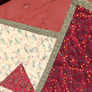 May include: A close-up of a red, green, and cream quilted fabric with a star-shaped design. The fabric is quilted with a pattern of holly leaves and berries.