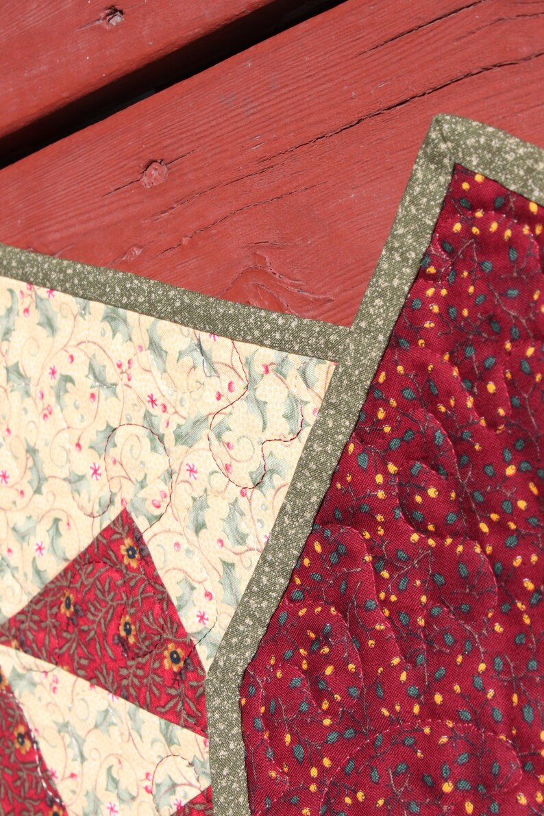 May include: A close-up of a red, green, and cream quilted fabric with a star-shaped design. The fabric is quilted with a pattern of holly leaves and berries.